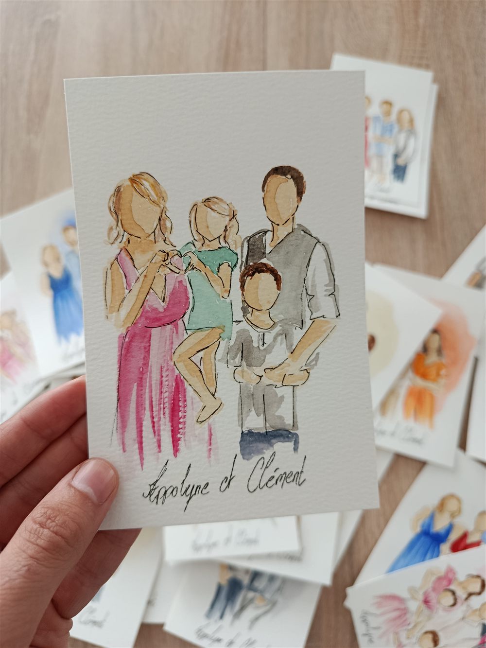 Mini portrait aquarelle d'une famille réalisé en direct lors d'un événement, tenu à la main avec d'autres cartes en arrière-plan