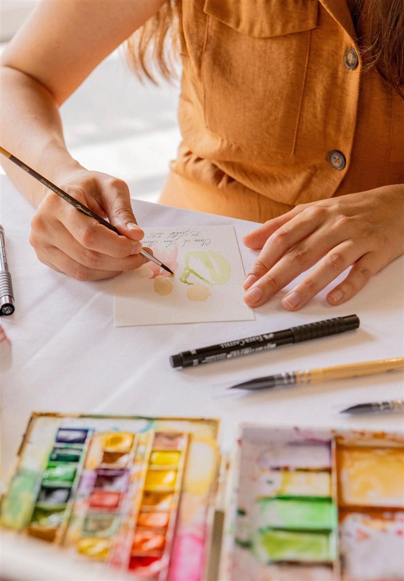 Artiste de L'Atelier B en train de peindre à l'aquarelle, gros plan sur les mains, le pinceau et la palette de couleurs